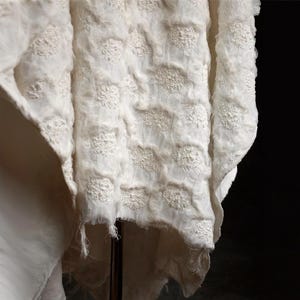 Könnte beinhalten: Cremefarbener Stoff mit einem strukturierten, gepunkteten Muster. Das Material scheint geschichtet zu sein, mit einer transparenten Überlagerung, die detaillierte, erhabene, blütenartige Designs zeigt. Die Kanten sind leicht ausgefranst, was eine zarte, handgefertigte Qualität verleiht.