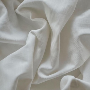 Könnte beinhalten: Nahaufnahme eines weißen Stoffes mit gerippter Textur. Der Stoff ist drapiert und gefaltet, wodurch weiche Schatten und Lichteffekte entstehen. Das Material wirkt weich und fließend, was auf ein Kleidungsstück oder Textil hindeutet.