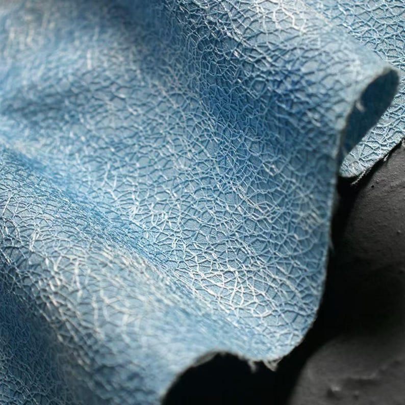 Op de afbeelding: Close-up van een lichtblauwe stof met een textuur en een netwerk van witte, webachtige patronen. De stof is gevouwen, waardoor de gelaagde textuur en subtiele kleurvariaties zichtbaar zijn. Het materiaal lijkt zacht en soepel.