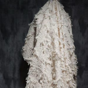 May include: A draped, off-white textile with a heavily textured surface. The fabric is covered in short, frayed strands, creating a shaggy, layered effect. The background is a dark, mottled gray.