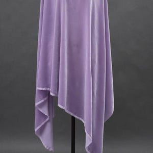 May include: A draped, lavender-colored fabric with a soft, velvety texture. The fabric is hanging from a black stand, with a folded, asymmetrical design. The edges are finished with a lighter-colored trim, creating a subtle contrast.