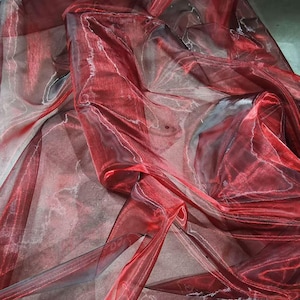 May include: Close-up of a flowing, sheer fabric in shades of red and burgundy. The fabric has a glossy sheen and a marbled appearance, with lighter veins running through the darker colors. The material is draped and folded, creating a sense of movement and texture.