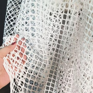 Könnte beinhalten: Nahaufnahme eines weißen Stoffes mit offenem Gewebe und einem gitterartigen Muster. Der Stoff wird hochgehalten, wodurch seine Textur und die Art und Weise, wie Licht durch die offenen Räume scheint, sichtbar werden. Das Material scheint weich und flexibel zu sein.