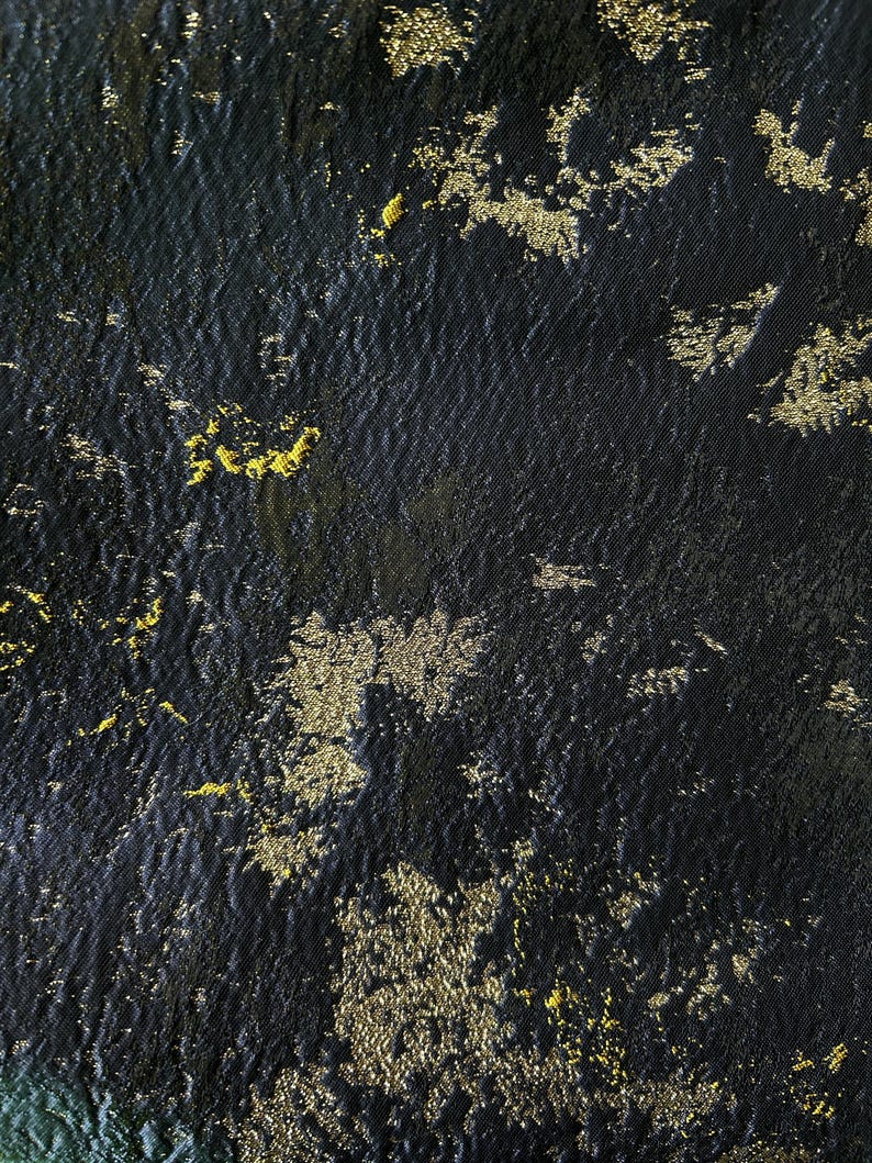 May include: Close-up of a textured black fabric with gold metallic accents. The gold elements are irregularly shaped and scattered across the surface, creating a shimmering effect. The fabric appears to have a subtle sheen.