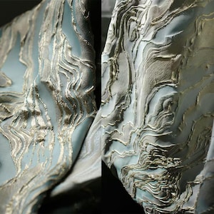 May include: Close-up of a luxurious fabric with a flowing, marbled design. The fabric features a light blue base with intricate gold-colored, raised patterns. The texture appears soft and the design is reminiscent of flowing water or stone.