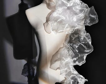 May include: A white dress form draped with a flowing, sheer, ruffled fabric. The fabric cascades down the form, creating a layered effect. The dress form is set against a dark background, with a shadow of another form visible.