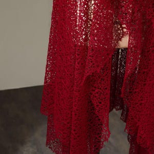 May include: A flowing, deep red lace shawl with a delicate, open weave. The fabric drapes in soft folds, creating a textured and elegant appearance. The shawl's intricate design and rich color suggest a formal or special occasion garment.