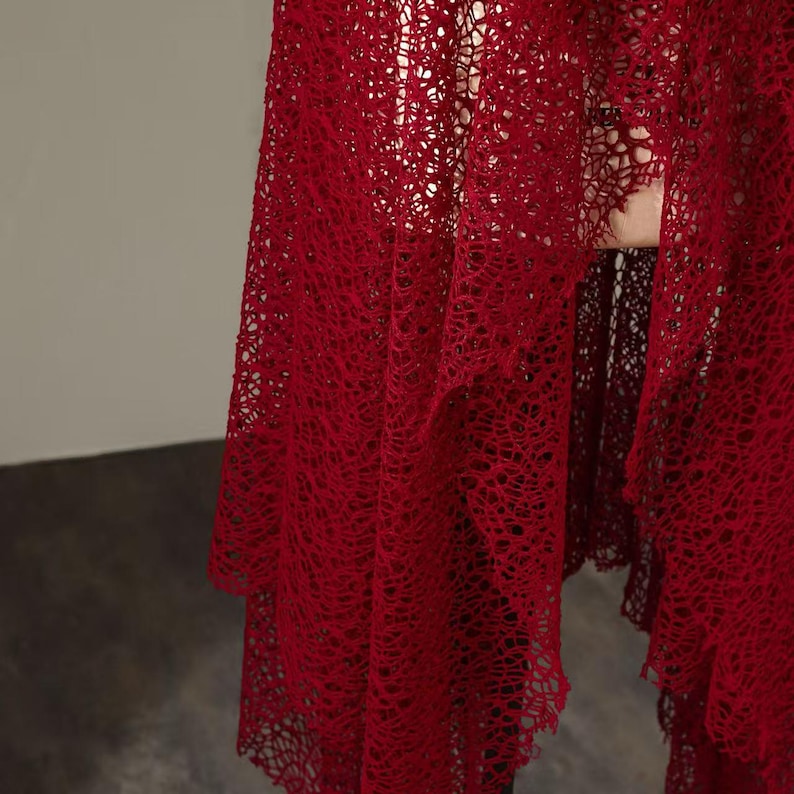 May include: A flowing, deep red lace shawl with a delicate, open weave. The fabric drapes in soft folds, creating a textured and elegant appearance. The shawl's intricate design and rich color suggest a formal or special occasion garment.
