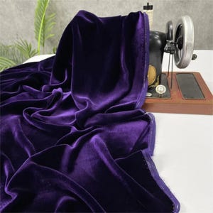 May include: A bolt of luxurious, deep purple velvet fabric draped over a vintage black sewing machine. The fabric has a soft, lustrous sheen and is gathered in elegant folds. The sewing machine is set on a wooden base.