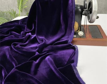 May include: A length of rich, deep purple velvet fabric draped over a vintage black sewing machine. The fabric has a soft, lustrous sheen and is gathered in elegant folds. The sewing machine is set on a wooden base.