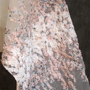 Puede incluir: Primer plano de una tela transparente con un diseño floral. La tela está adornada con flores texturizadas en tonos blanco, rosa pálido y azul claro. El delicado material es probablemente para ropa o fines decorativos.