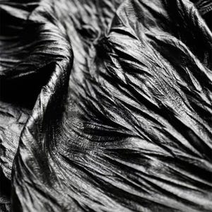 May include: Close-up of a textured, black fabric with a rippled, flowing appearance. The material has a shiny, almost metallic sheen, with light reflecting off the folds and creases. The fabric's surface is detailed with fine lines and subtle variations in tone.