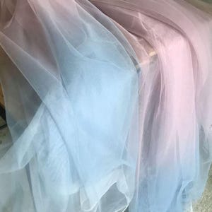 Op de afbeelding: Close-up van een transparante stof met een kleurovergang. De stof verloopt van lichtblauw naar zachtroze. Het materiaal lijkt tule of een vergelijkbaar lichtgewicht textiel te zijn, waardoor een delicaat, luchtig effect ontstaat.
