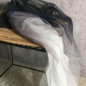Puede incluir: Capas de tela transparente en tonos negro, gris y blanco están drapeadas sobre una superficie de madera. La tela parece ligera y delicada, con un sutil efecto degradado. El material es probablemente tul o una red similar.