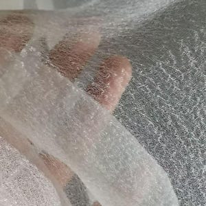 Puede incluir: Primer plano de una tela blanca transparente con un tejido delicado y abierto. La tela parece ligeramente translúcida, con un brillo sutil. La textura es suave y aireada, lo que sugiere un material ligero.