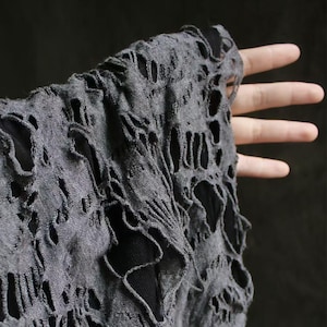May include: Close-up of a dark gray, distressed fabric with numerous cut-out patterns. The fabric appears to be a lightweight knit, with a textured surface. The design features irregular, open areas, creating a unique, worn aesthetic. The fabric is held up by a hand.