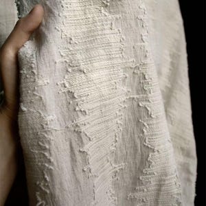 May include: Close-up of off-white fabric with a textured, woven design. The fabric has a slightly distressed appearance with visible stitching and frayed edges. The material is draped, showcasing the texture and weave of the textile.