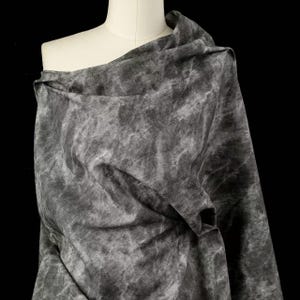 May include: A draped fabric in shades of gray, with a marbled or tie-dye effect. The fabric is draped over a mannequin, showcasing its texture and flow. The background is black, highlighting the fabric's details.