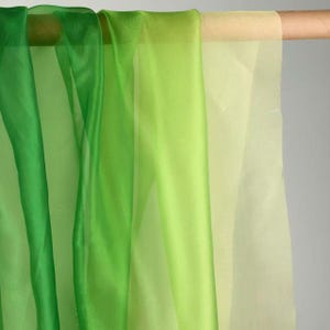 May include: Sheer fabric in shades of green and yellow hangs from a wooden rod. The fabric is layered, creating a gradient effect from deep green to pale yellow. The material appears lightweight and translucent, suitable for curtains or decorative purposes.