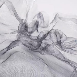 May include: Close-up of a flowing, sheer fabric with a delicate mesh pattern. The fabric is a dark gray color, with areas of lighter gray and white, creating a sense of depth and movement. The material is draped and folded, suggesting a textile or decorative element.