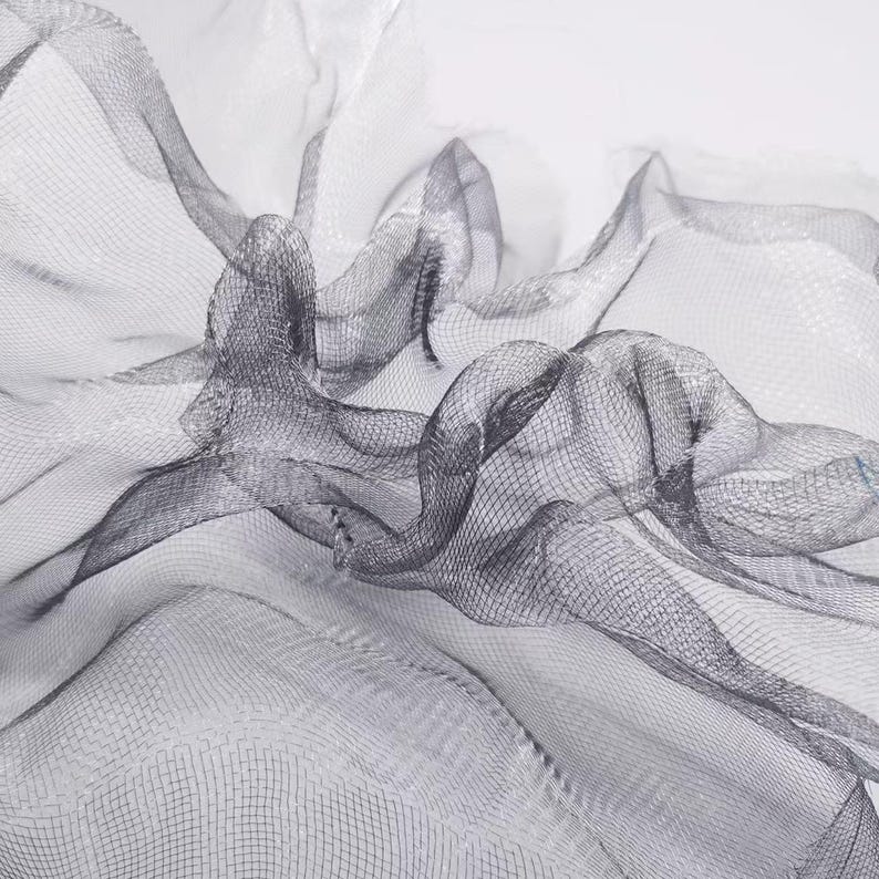 May include: Close-up of a flowing, sheer fabric with a delicate mesh pattern. The fabric is a dark gray color, with areas of lighter gray and white, creating a sense of depth and movement. The material is draped and folded, suggesting a textile or decorative element.