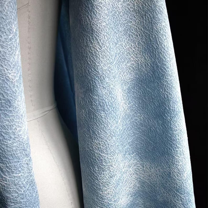Op de afbeelding: Close-up van een lichtblauwe stof met een getextureerd, webachtig patroon. De stof is over een mannequin gedrapeerd, waarbij de textuur een subtiel, ingewikkeld ontwerp cre&euml;ert. De kleur is een zacht, gedempt blauw en de stof lijkt licht van gewicht.