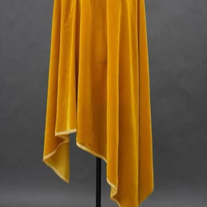 May include: Mustard yellow velvet fabric draped over a black stand. The fabric has a soft, luxurious texture and is trimmed with a lighter gold edge. The folds and draping create a sense of movement and depth.