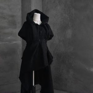 May include: A black hooded cape and matching pants set. The cape features a draped design with a hood and button closure. The pants have a layered, asymmetrical hem. The outfit is displayed on a mannequin against a gray background.