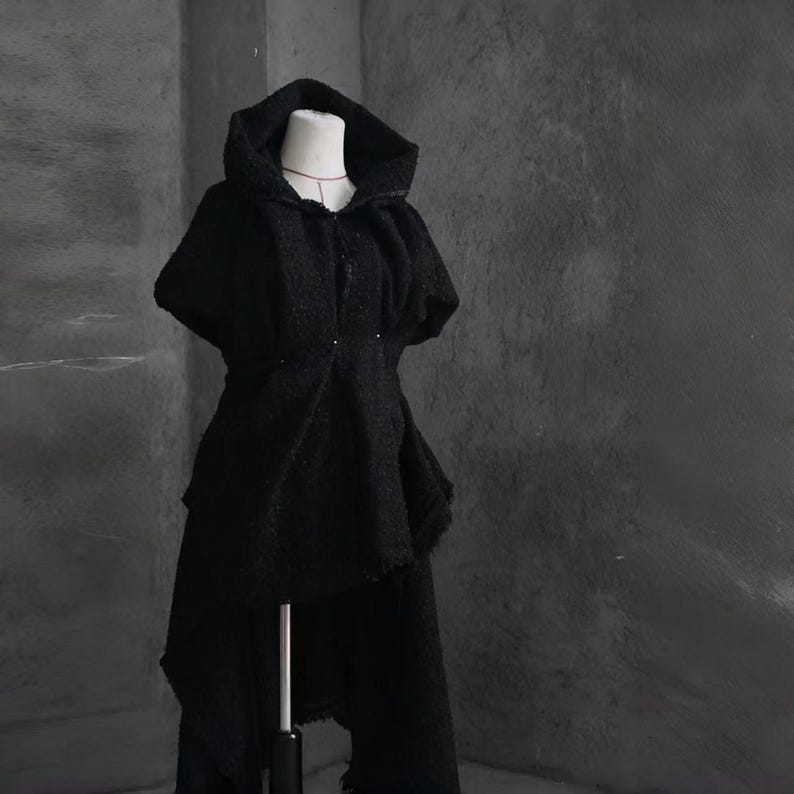 May include: A black hooded cape and matching pants set. The cape features a draped design with a hood and button closure. The pants have a layered, asymmetrical hem. The outfit is displayed on a mannequin against a gray background.