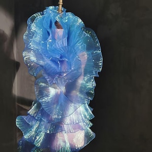 Può includere: Un abito blu a strati con un design a balze. L'abito è realizzato in un tessuto cangiante e traslucido, creando un look fluido ed etereo. L'abito è esposto su un manichino, mettendo in mostra i suoi intricati dettagli e la sua forma.