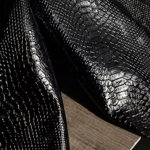 Könnte beinhalten: Nahaufnahme von schwarzem, strukturiertem Leder mit glänzendem Finish. Das Leder hat ein schuppenartiges Muster, und die Falten erzeugen Schatten, die die Textur hervorheben. Das Material scheint hochwertig und langlebig zu sein.