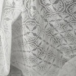 May include: Close-up of a sheer white fabric with a repeating pattern of stylized floral and geometric shapes. The fabric is draped, creating soft folds and shadows. The design is made of small white dots.