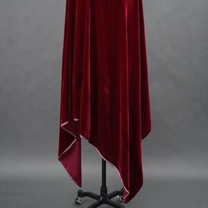 May include: A rich, deep red velvet cloak with white edging. The cloak is displayed on a black metal stand with a wheeled base, against a grey background. The fabric appears soft and luxurious, with a flowing drape.