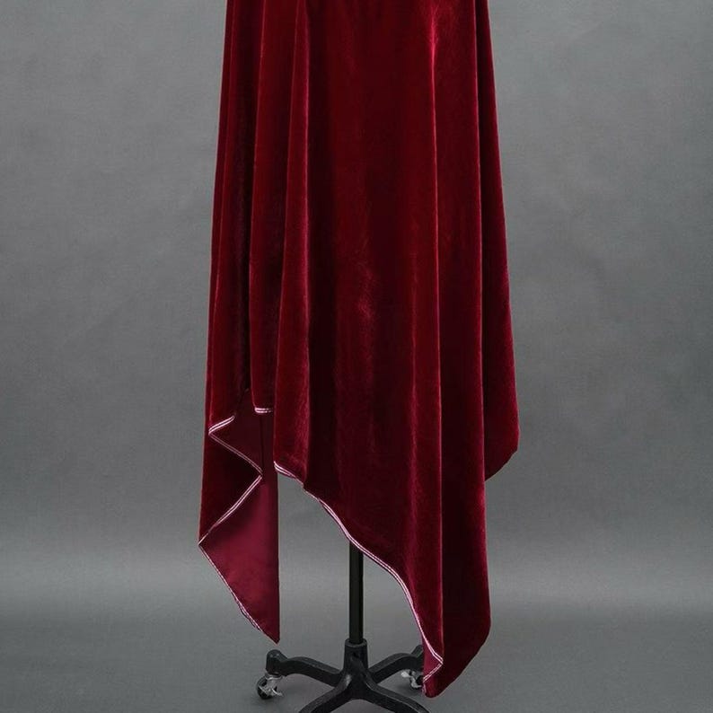 May include: A rich, deep red velvet cloak with white edging. The cloak is displayed on a black metal stand with a wheeled base, against a grey background. The fabric appears soft and luxurious, with a flowing drape.