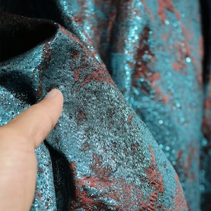 Può includere: Primo piano di un tessuto broccato verde acqua e rosso. Il tessuto ha una superficie strutturata con un effetto scintillante. Il motivo presenta disegni floreali rossi su sfondo verde acqua. Il tessuto è piegato, rivelando l'interno.