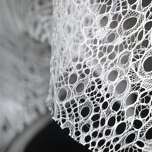 May include: Close-up of a white, open-weave fabric with a delicate, lacy pattern. The fabric has a repeating design of irregular, oval-shaped holes connected by thin threads, creating a textured surface. The material is likely used for decorative purposes.