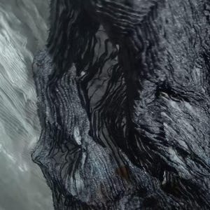 May include: Close-up of black fabric with a textured, layered appearance. The fabric has a flowing, rippled design with varying shades of black and gray, creating a sense of depth and movement. The material appears to be lightweight and possibly sheer.