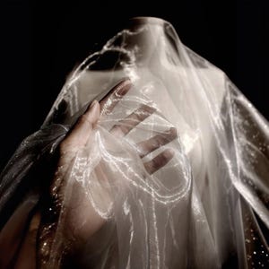 May include: A hand holds a sheer, shimmering white fabric, possibly organza or a similar material. The fabric is draped over a mannequin, with light catching the delicate texture and creating a luminous effect. The background is dark, emphasizing the fabric's ethereal quality.