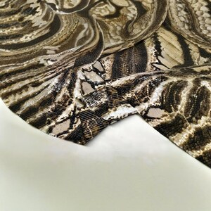 May include: Close-up of a fabric with a snakeskin pattern in shades of brown, tan, and black. The fabric has a textured appearance with subtle sequin embellishments along the edges, adding a touch of shimmer.