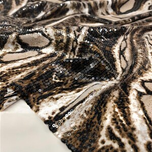 May include: A close-up shot of a fabric featuring a tiger stripe design in brown, beige, and white. The material is embellished with small, reflective sequins. The fabric is folded, revealing its texture and how it drapes.