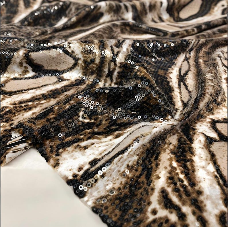 May include: A close-up shot of a fabric featuring a tiger stripe design in brown, beige, and white. The material is embellished with small, reflective sequins. The fabric is folded, revealing its texture and how it drapes.