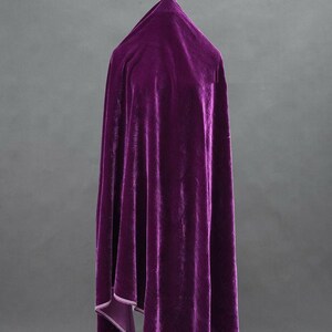 May include: A draped, deep purple velvet fabric. The material is gathered at the top and falls in soft folds. The edges of the fabric are lined with a lighter purple material. The background is a solid grey.