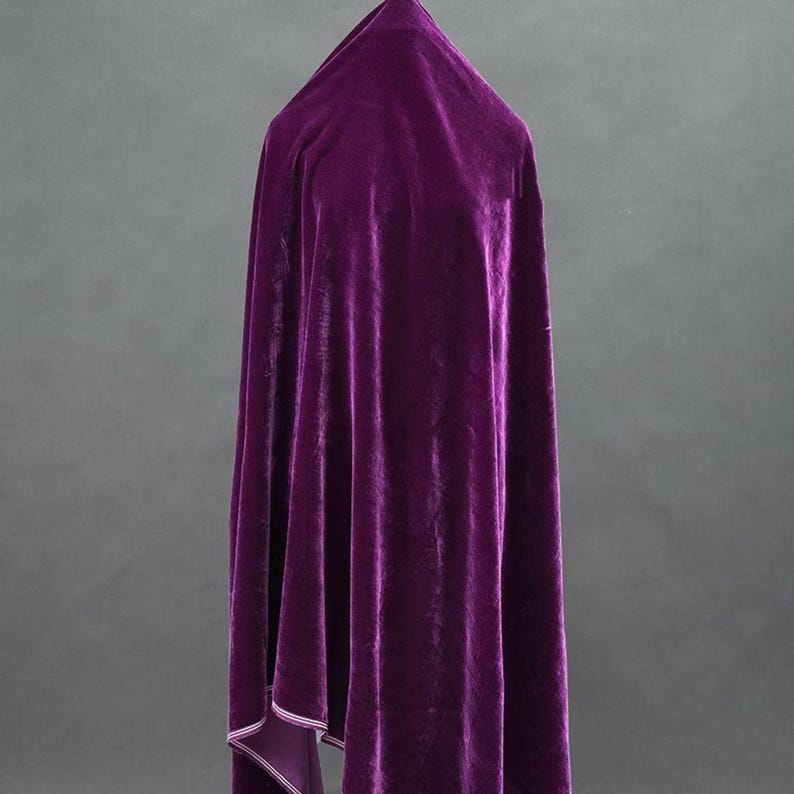 May include: A draped, deep purple velvet fabric. The material is gathered at the top and falls in soft folds. The edges of the fabric are lined with a lighter purple material. The background is a solid grey.