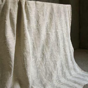 May include: Off-white textured fabric with a vertical striped pattern. The fabric is draped over a surface, creating soft folds and shadows. The material appears to be thick and woven, with a slightly rough texture.