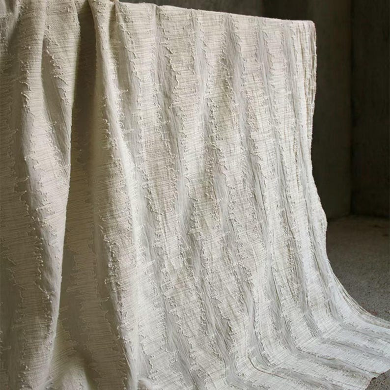 May include: Off-white textured fabric with a vertical striped pattern. The fabric is draped over a surface, creating soft folds and shadows. The material appears to be thick and woven, with a slightly rough texture.