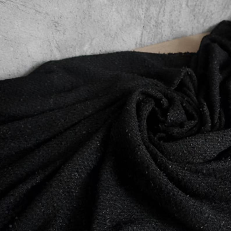 May include: A close-up shot of a dark, textured fabric with a woven appearance. The fabric is a deep black color, with subtle variations in tone and texture. The material is draped and folded, creating shadows and highlights.