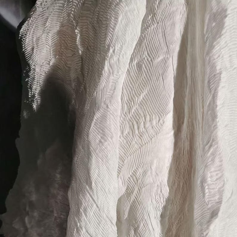May include: Close-up of white fabric with a textured, rippled surface. The fabric has a flowing, draped appearance, with visible diagonal lines and subtle shadows. The material appears soft and lightweight.