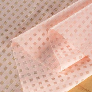 May include: A close-up of a folded, sheer fabric in a soft pink hue. The fabric has a repeating pattern of small, square, slightly darker pink shapes, creating a grid-like design. The folds and layers add depth and texture to the material.