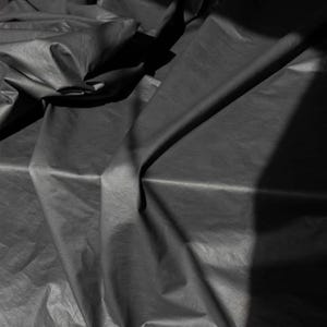 Op de afbeelding: Close-up van een donkergrijze stof met een gekreukte textuur. De stof is gevouwen en gedrapeerd, waardoor schaduwen en highlights ontstaan die de textuur benadrukken. De belichting is dramatisch, met een sterk contrast tussen lichte en donkere gebieden.