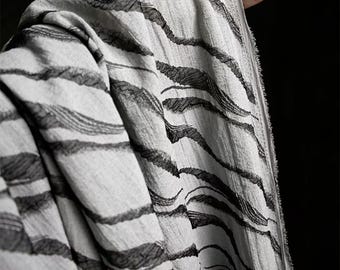 May include: Close-up of a light gray fabric with a black, wave-like pattern. The fabric is draped, showing the texture and design. The pattern resembles brushstrokes or abstract stripes, creating a flowing visual effect. The fabric appears soft and lightweight.